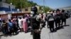 A Haitian National Police (PNH) officer looks on as Haitians stand behind him in the in the border of Malpasse, Haiti, March 17, 2020. 